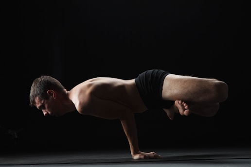 A person is performing an advanced yoga pose, balancing on their hands with their legs bent and tucked in. The background is dark, highlighting the subject's muscular form and intense focus.