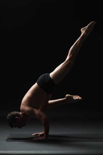 A person is performing a complex yoga balance pose on a mat. The individual is supporting their body on their hands, with their legs extended upward. The background is dark, emphasizing the strong posture and muscular definition.