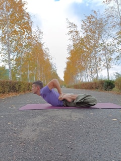 A person is performing a yoga pose on a mat, lying face down on a paved path surrounded by trees with autumn leaves. The trees form a symmetrical alley along the path, creating a picturesque and serene setting.