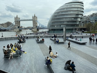 Several people are gathered on a modern plaza near a distinctive glass building with curved architecture. In the background, there is a large and iconic bridge spanning a river. People are seated on benches, walking, and standing around, creating a lively atmosphere.