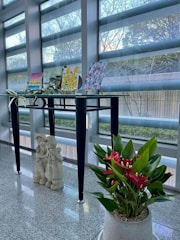 A modern glass table set near a large window displays several vibrant paintings propped on easels. Small green plants accompany the artwork, adding a touch of nature. Beneath the table, there is a pair of decorative stone statues depicting two children. A lush potted plant with vivid red flowers stands prominently in the foreground, adding color to the scene.