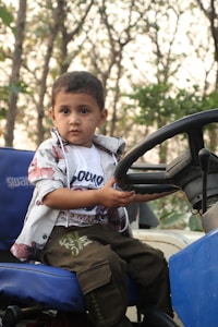 A young child, dressed in a white shirt with designs, a floral patterned jacket, and olive green pants, is sitting on the seat of a vehicle and holding onto the steering wheel. The background shows a forested area with trees and foliage.