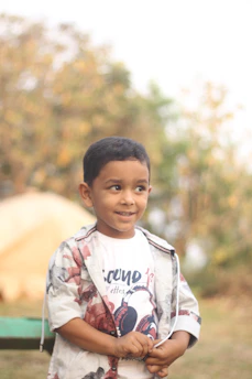 A cheerful young boy dressed in casual menswear, smiling outdoors.