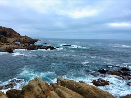 Waves crash against rugged rocks on the shoreline beneath a cloudy sky. The sea is a deep blue with patches of foamy white from the waves. Rocky cliffs extend into the distance on the left side, providing a dramatic backdrop.