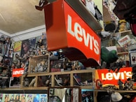 A cluttered room filled with various collectibles and memorabilia. Prominent Levi's signs are displayed among a dense arrangement of action figures, posters, and pop culture items such as Star Wars and music-related merchandise. Shelves are packed with an array of toys and models, creating a vibrant and chaotic visual atmosphere.