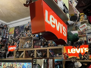 A cluttered room filled with various collectibles and memorabilia. Prominent Levi's signs are displayed among a dense arrangement of action figures, posters, and pop culture items such as Star Wars and music-related merchandise. Shelves are packed with an array of toys and models, creating a vibrant and chaotic visual atmosphere.