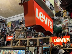 A cluttered room filled with various collectibles and memorabilia. Prominent Levi's signs are displayed among a dense arrangement of action figures, posters, and pop culture items such as Star Wars and music-related merchandise. Shelves are packed with an array of toys and models, creating a vibrant and chaotic visual atmosphere.