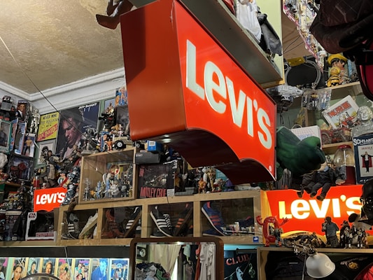 A cluttered room filled with various collectibles and memorabilia. Prominent Levi's signs are displayed among a dense arrangement of action figures, posters, and pop culture items such as Star Wars and music-related merchandise. Shelves are packed with an array of toys and models, creating a vibrant and chaotic visual atmosphere.