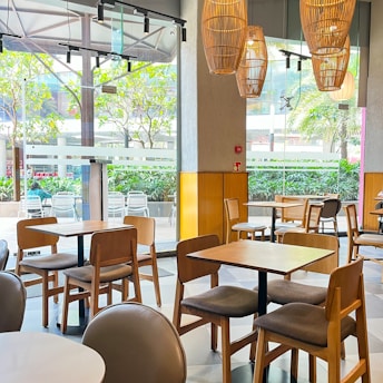 A cozy indoor café setting with several wooden tables and chairs. Large windows allow natural light to flood the space, highlighting the wooden accents and stylish wicker pendant lights hanging from the ceiling. Outside, greenery and seating areas are visible through the glass.