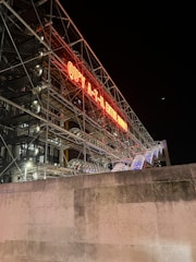 A building structure at night with an exposed industrial style featuring metal frames and glass. A prominent neon sign in French reads 'QU'Y A-T-IL ENTRE NOUS?' There are visible tubular escalators with lights along the structure.