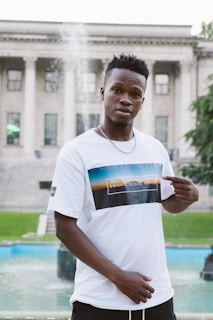 A man stands outdoors in front of a historic building with large columns. He is wearing a white T-shirt with a graphic and text that reads 'Swahili to the World'. There is a fountain in the background, and he is pointing to his T-shirt with both hands.