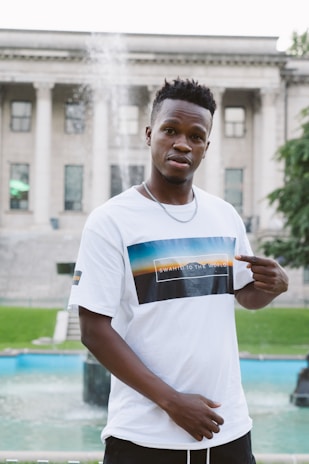 A man stands outdoors in front of a historic building with large columns. He is wearing a white T-shirt with a graphic and text that reads 'Swahili to the World'. There is a fountain in the background, and he is pointing to his T-shirt with both hands.
