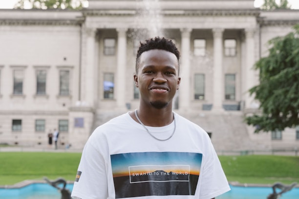 A person wearing a white t-shirt with a graphic print stands in front of a classical building with columns. A green lawn and a pool can be seen in the background, along with a few blurred figures.