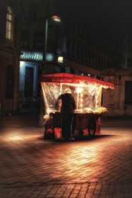 A lone street vendor stands at a brightly lit food cart on a cobblestone street at night. The vendor's back is turned, and the warm glow from the cart illuminates the immediate area in contrast to the dark surroundings. The scene appears quiet and atmospheric, with dimly visible buildings in the background.