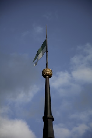 A tall flagpole topped with a decorative gold sphere, bearing a green and white striped flag fluttering gently. The background consists of a sky with dispersed clouds, suggesting a clear day.