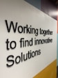 Black 3D text displays the phrase 'Working together to find innovative Solutions' against a geometric background with white, yellow, light blue, and dark blue areas.