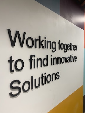 Black 3D text displays the phrase 'Working together to find innovative Solutions' against a geometric background with white, yellow, light blue, and dark blue areas.