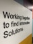 Black 3D text displays the phrase 'Working together to find innovative Solutions' against a geometric background with white, yellow, light blue, and dark blue areas.
