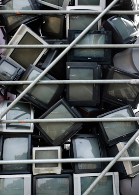 A stack of old CRT televisions is piled behind metal bars, presenting a chaotic and cluttered scene. The televisions vary in size and color, predominantly composed of black and gray frames, some showing reflections on their screens.