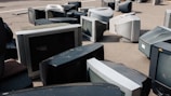 A collection of old, bulky CRT televisions scattered across an outdoor pavement. The televisions vary in size and are arranged haphazardly, some lying on their sides while others are upright. The scene suggests abandonment or disposal, as the screens appear dusty and outdated.