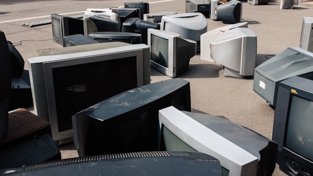 A collection of old, bulky CRT televisions scattered across an outdoor pavement. The televisions vary in size and are arranged haphazardly, some lying on their sides while others are upright. The scene suggests abandonment or disposal, as the screens appear dusty and outdated.