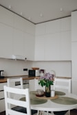 A modern kitchen with white cabinets and a wooden countertop. A small, round dining table with white chairs, adorned with a vase of purple flowers. Various kitchen appliances, such as a toaster and microwave, are visible on the counter.