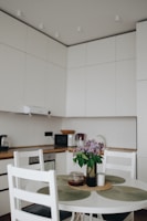 A modern kitchen with white cabinets and a wooden countertop. A small, round dining table with white chairs, adorned with a vase of purple flowers. Various kitchen appliances, such as a toaster and microwave, are visible on the counter.