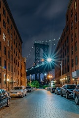 A nighttime street scene with a view of the Manhattan Bridge framed between two rows of brick buildings. Cars are parked along both sides of the cobblestone street, and the bridge is brightly illuminated with lights, casting a vibrant glow. The sky is dark, suggesting it's either evening or night.