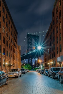 A nighttime street scene with a view of the Manhattan Bridge framed between two rows of brick buildings. Cars are parked along both sides of the cobblestone street, and the bridge is brightly illuminated with lights, casting a vibrant glow. The sky is dark, suggesting it's either evening or night.