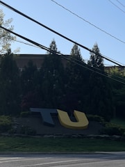Large, stylized letters 'T' and 'U' are positioned on a landscaped area near a road. In the background, there are tall evergreen trees and part of a brick building. Power lines cross the top portion of the image under a clear blue sky.