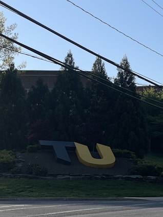 Large, stylized letters 'T' and 'U' are positioned on a landscaped area near a road. In the background, there are tall evergreen trees and part of a brick building. Power lines cross the top portion of the image under a clear blue sky.