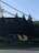 Large, stylized letters 'T' and 'U' are positioned on a landscaped area near a road. In the background, there are tall evergreen trees and part of a brick building. Power lines cross the top portion of the image under a clear blue sky.