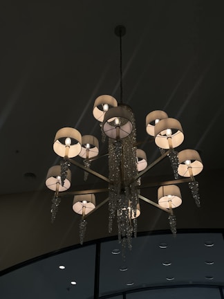A luxurious chandelier with multiple lights, featuring elegantly decorated lamp shades and crystal ornaments hanging from each arm. The background shows a dark ceiling with high-placed circular fixtures.