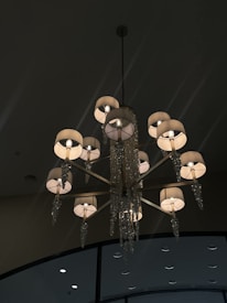 A luxurious chandelier with multiple lights, featuring elegantly decorated lamp shades and crystal ornaments hanging from each arm. The background shows a dark ceiling with high-placed circular fixtures.