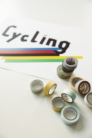 A set of colorful tape rolls is arranged on a light-colored surface beside a white paper featuring the word 'Cycling' in bold, black letters. Below the text, there are horizontal stripes in blue, red, black, yellow, and green.