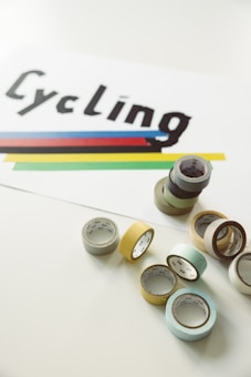 A set of colorful tape rolls is arranged on a light-colored surface beside a white paper featuring the word 'Cycling' in bold, black letters. Below the text, there are horizontal stripes in blue, red, black, yellow, and green.