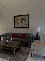 A cozy living room setting with a gray tufted sofa adorned with two red cushions. A wooden coffee table sits in front of the sofa, holding a bouquet of colorful flowers. A map in a black frame hangs on the wall above the sofa. A patterned armchair with light upholstery is positioned next to the sofa. There are two table lamps with white shades on either side of the room, and the floor is covered with a multicolored rug.