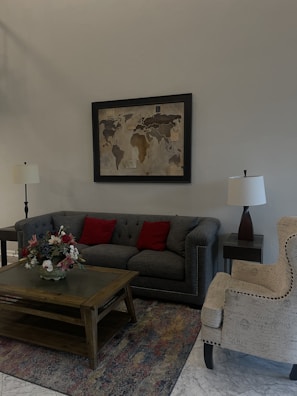 A cozy living room setting with a gray tufted sofa adorned with two red cushions. A wooden coffee table sits in front of the sofa, holding a bouquet of colorful flowers. A map in a black frame hangs on the wall above the sofa. A patterned armchair with light upholstery is positioned next to the sofa. There are two table lamps with white shades on either side of the room, and the floor is covered with a multicolored rug.