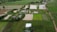 An aerial view of an agricultural landscape with rectangular plots of land, some filled with water indicating rice paddies. There are various shades of green vegetation and a few scattered small structures. A dirt road runs through the scene, with trees and bushes lining the plots.