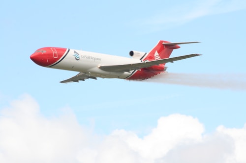 A brightly colored aircraft with red and white markings is flying in a clear blue sky. The plane has the words 'Oil Spill Response' on its side and is releasing a mist or spray from its underside.