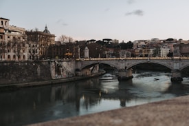 A picturesque scene featuring a stone bridge crossing a river, with historic architecture lining both sides. Trees with bare branches suggest it is autumn or winter, while the soft lighting creates a calm and serene atmosphere.