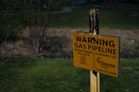 A yellow warning sign is posted in a grassy area near a wooded landscape. It cautions about an underground gas pipeline, providing emergency contact information and instructions to call before digging.