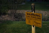 A yellow warning sign is posted in a grassy area near a wooded landscape. It cautions about an underground gas pipeline, providing emergency contact information and instructions to call before digging.
