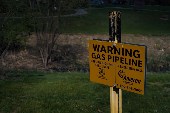A yellow warning sign is posted in a grassy area near a wooded landscape. It cautions about an underground gas pipeline, providing emergency contact information and instructions to call before digging.