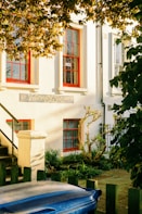 A quaint veterinary clinic with a classic exterior, featuring white walls and bright red-framed windows. Sunlight filters through the leaves of a tree, casting dappled shadows on the building. A small garden with shrubs and plants lines the entrance, while a blue trash bin and a wooden fence are visible in the foreground.