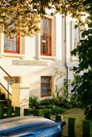 A quaint veterinary clinic with a classic exterior, featuring white walls and bright red-framed windows. Sunlight filters through the leaves of a tree, casting dappled shadows on the building. A small garden with shrubs and plants lines the entrance, while a blue trash bin and a wooden fence are visible in the foreground.