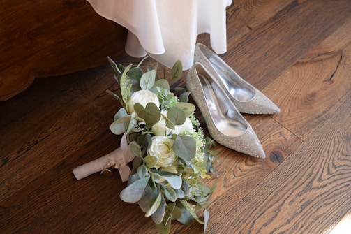 A pair of elegant, sparkly high-heeled shoes are placed on a wooden floor next to a lush bridal bouquet with white roses and greenery. The bottom of a flowing white dress is visible above the shoes.