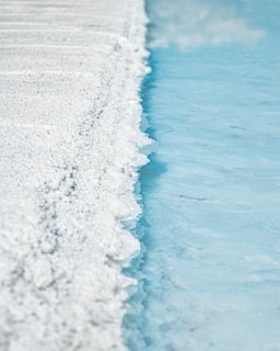 A textured surface of white crystalline salt is adjacent to a calm blue body of water. The salt forms a rough, uneven edge against the smooth water, creating a stark contrast between the two elements.