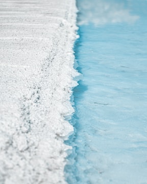A textured surface of white crystalline salt is adjacent to a calm blue body of water. The salt forms a rough, uneven edge against the smooth water, creating a stark contrast between the two elements.