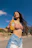 A smiling woman holding a chilled bottle of Frutyka mango juice against a sunny outdoor backdrop.